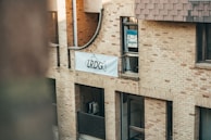 An exterior view of a multi-story brick building with a sign displaying the letters 'LRDG' on a white banner. The facade includes arched architectural features and windows, with some windows having visible objects and posters inside. The scene is lit by natural light, creating a warm and calm atmosphere.
