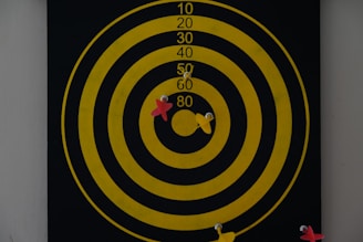 A dartboard with concentric yellow circles on a black background displays several darts. Two red and two yellow darts are embedded in the board around the center circle, indicating various attempts at hitting the bullseye.