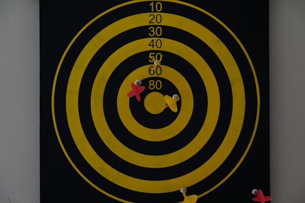 A dartboard with concentric yellow circles on a black background displays several darts. Two red and two yellow darts are embedded in the board around the center circle, indicating various attempts at hitting the bullseye.