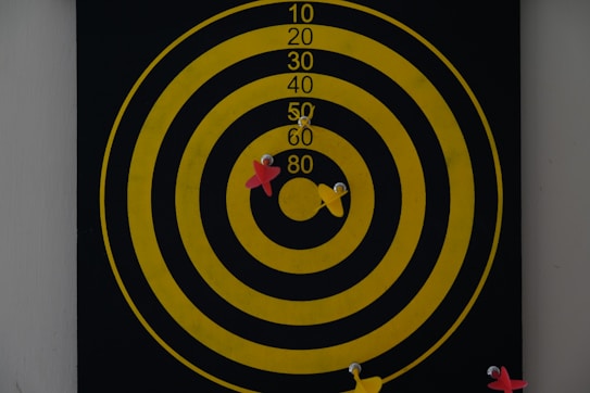 A dartboard with concentric yellow circles on a black background displays several darts. Two red and two yellow darts are embedded in the board around the center circle, indicating various attempts at hitting the bullseye.