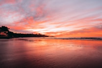 The image displays a serene beach at sunset with a vibrant sky painted in shades of orange, pink, and purple. The calm ocean waves gently lap against the shore, reflecting the colorful hues of the sky. Silhouettes of distant trees and structures are visible along the shoreline.