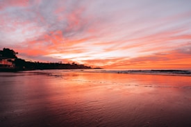 The image displays a serene beach at sunset with a vibrant sky painted in shades of orange, pink, and purple. The calm ocean waves gently lap against the shore, reflecting the colorful hues of the sky. Silhouettes of distant trees and structures are visible along the shoreline.