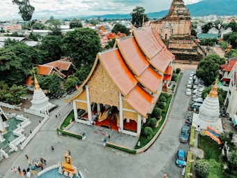 Aerial view of a traditional temple complex featuring ornate roofs with golden and red accents. Surrounding the temple are lush green trees and smaller structures, including white stupas with gold tips. People are scattered across the temple grounds, and several cars are parked nearby. The backdrop includes distant hills and city structures.