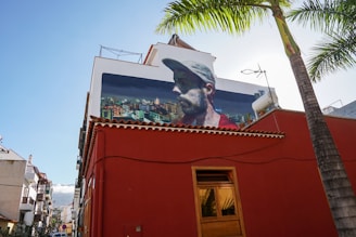 A red building features a large mural on an upper wall, depicting a profile of a man in a cap with an urban skyline in the background. Palm trees and blue sky can be seen nearby, and multiple buildings are visible along the street.
