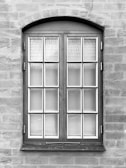 A symmetrical, rectangular window framed with dark wood is set into a textured stone wall. The window consists of two panels, each containing multiple small glass panes separated by white muntins. The surface of the wall appears uneven with visible brush strokes or trowel marks.
