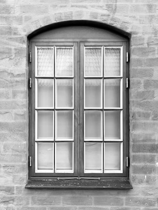 A symmetrical, rectangular window framed with dark wood is set into a textured stone wall. The window consists of two panels, each containing multiple small glass panes separated by white muntins. The surface of the wall appears uneven with visible brush strokes or trowel marks.