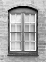 A symmetrical, rectangular window framed with dark wood is set into a textured stone wall. The window consists of two panels, each containing multiple small glass panes separated by white muntins. The surface of the wall appears uneven with visible brush strokes or trowel marks.