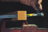 A hand appears to be working with a piece of equipment that holds a rectangular block of wood in place. The block is clamped by a metal vise, and there are yellow and green paint marks on the metal. Fine particles are visible around the wooden block, possibly indicating sanding or cutting.