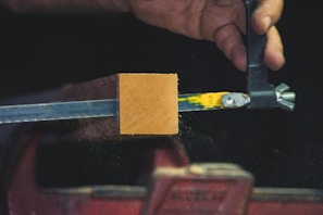 A hand appears to be working with a piece of equipment that holds a rectangular block of wood in place. The block is clamped by a metal vise, and there are yellow and green paint marks on the metal. Fine particles are visible around the wooden block, possibly indicating sanding or cutting.