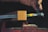 A hand appears to be working with a piece of equipment that holds a rectangular block of wood in place. The block is clamped by a metal vise, and there are yellow and green paint marks on the metal. Fine particles are visible around the wooden block, possibly indicating sanding or cutting.