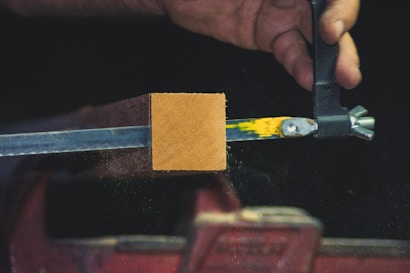 A hand appears to be working with a piece of equipment that holds a rectangular block of wood in place. The block is clamped by a metal vise, and there are yellow and green paint marks on the metal. Fine particles are visible around the wooden block, possibly indicating sanding or cutting.