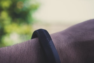 A close-up of a person's wrist wearing a black, textured fitness band. The background is blurred with greenish hues, suggesting an outdoor setting.