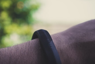 A close-up of a person's wrist wearing a black, textured fitness band. The background is blurred with greenish hues, suggesting an outdoor setting.