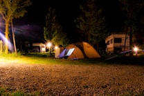 A nighttime camping scene features a well-lit tent situated on a grassy area with gravel underfoot. In the background, several trees and two RVs are visible under the dark sky. Bright artificial lights illuminate the surroundings, casting warm glows and light flares.