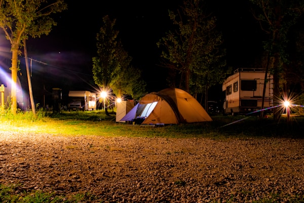 A nighttime camping scene features a well-lit tent situated on a grassy area with gravel underfoot. In the background, several trees and two RVs are visible under the dark sky. Bright artificial lights illuminate the surroundings, casting warm glows and light flares.