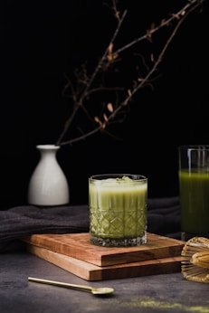 a glass of green juice next to a wooden cutting board