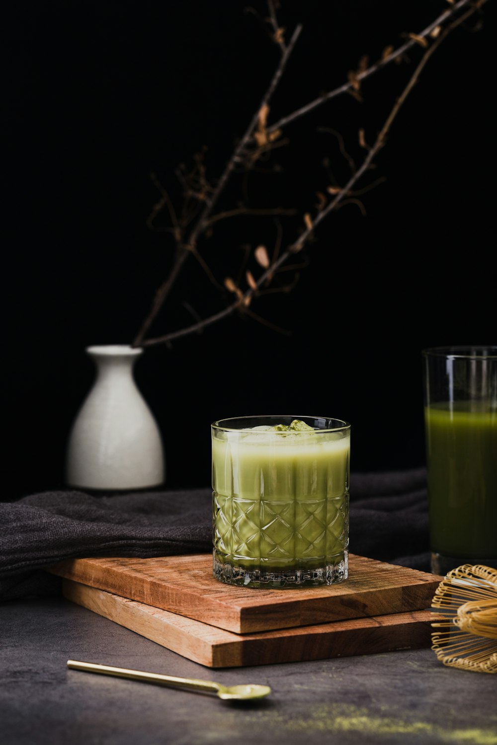 a glass of green juice next to a wooden cutting board