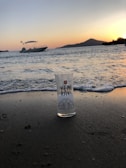 A glass with 'Yeni Rakı' branding stands on a sandy beach as gentle waves approach. In the background, a boat is anchored near the shore, with a silhouette of a mountain and trees visible. The sky features a gradient of colors from a setting sun.