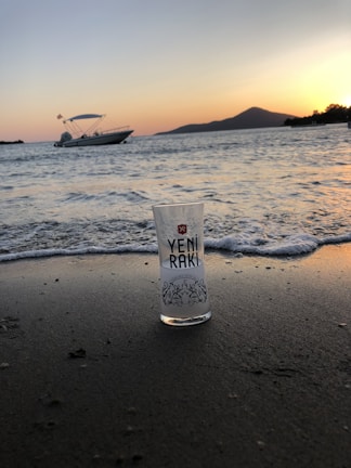A glass with 'Yeni Rakı' branding stands on a sandy beach as gentle waves approach. In the background, a boat is anchored near the shore, with a silhouette of a mountain and trees visible. The sky features a gradient of colors from a setting sun.