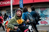 A person in a hoodie and hat is in a wheelchair on a busy street, with a couple of people walking by and covered storefronts behind showing graffiti. They carry a Chilean flag and other colorful items on the wheelchair.
