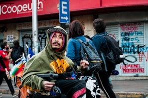 A person in a hoodie and hat is in a wheelchair on a busy street, with a couple of people walking by and covered storefronts behind showing graffiti. They carry a Chilean flag and other colorful items on the wheelchair.