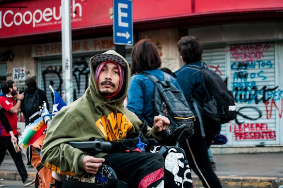 A person in a hoodie and hat is in a wheelchair on a busy street, with a couple of people walking by and covered storefronts behind showing graffiti. They carry a Chilean flag and other colorful items on the wheelchair.