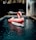 A large pink flamingo-shaped inflatable float is situated in a small, rectangular swimming pool. In front of the float is a white surfboard partially submerged in the water. The setting includes a wooden deck surrounding the pool, with a lounge chair and a ladder resting against a textured wall on the right. The ambiance appears relaxed and tropical, with some foliage visible in the background.