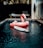 A large pink flamingo-shaped inflatable float is situated in a small, rectangular swimming pool. In front of the float is a white surfboard partially submerged in the water. The setting includes a wooden deck surrounding the pool, with a lounge chair and a ladder resting against a textured wall on the right. The ambiance appears relaxed and tropical, with some foliage visible in the background.