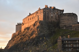 A historic castle stands majestically atop a rocky hill, bathed in warm golden light from the setting sun. The structure features stone walls, turrets, and a circular bastion. The sky is a gentle gradient of orange and blue, enhancing the dramatic and timeless aura of the scene.