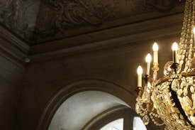 An ornate chandelier hangs from a ceiling adorned with intricate carvings. The chandelier features multiple candle-like lights and numerous crystal decorations that catch the light beautifully, casting a warm glow onto the arched window and detailed ceiling.