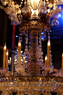 An ornate crystal chandelier with lit candles, decorated with hanging glass beads and intricate metalwork details. The setting is illuminated by warm candlelight and hints of blue light, creating a luxurious ambiance.