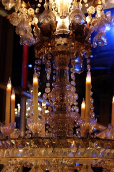 An ornate crystal chandelier with lit candles, decorated with hanging glass beads and intricate metalwork details. The setting is illuminated by warm candlelight and hints of blue light, creating a luxurious ambiance.
