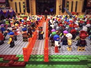 A large assembly of Lego minifigures arranged on Lego baseplates, with diverse outfits and helmets. The figures are gathered as if attending an event or festival, surrounded by orange barriers. In the background, there are Lego structures resembling buildings or a stage with intricate details.