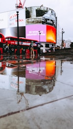 A rainy day scene featuring a wet pavement reflecting bright digital advertisements displayed on large screens. Two red double-decker buses, iconic to London, are parked nearby. Several pedestrians with umbrellas are visible, adding to the urban atmosphere.