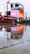 A rainy day scene featuring a wet pavement reflecting bright digital advertisements displayed on large screens. Two red double-decker buses, iconic to London, are parked nearby. Several pedestrians with umbrellas are visible, adding to the urban atmosphere.
