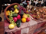 A basket overflowing with a variety of fruits including apples, grapes, and citrus fruits, placed on a patterned cloth-covered table. Autumn leaves, pine cones, and ornamental berries are arranged beside the basket.