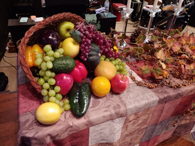A basket overflowing with a variety of fruits including apples, grapes, and citrus fruits, placed on a patterned cloth-covered table. Autumn leaves, pine cones, and ornamental berries are arranged beside the basket.
