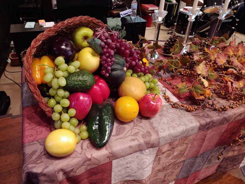 A basket overflowing with a variety of fruits including apples, grapes, and citrus fruits, placed on a patterned cloth-covered table. Autumn leaves, pine cones, and ornamental berries are arranged beside the basket.
