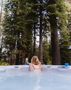 A person with blonde hair is relaxing in an outdoor hot tub surrounded by tall evergreen trees. They are facing away from the camera, wearing a red swimsuit while reaching for a drink placed on the edge of the tub. The scene conveys a sense of tranquility and nature.
