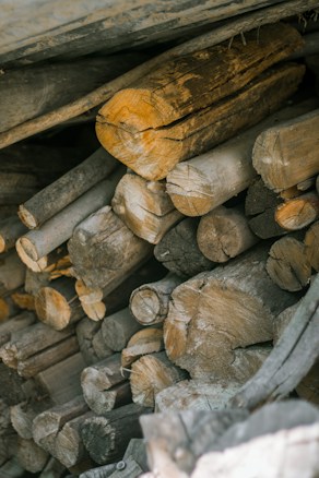 A stack of chopped firewood arranged in a pile. The logs vary in size and color, showing their natural texture and grain. They appear dry and seasoned, suitable for burning. The image conveys a sense of rustic simplicity and practical utility.