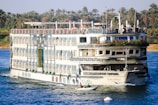 A large, multi-deck river cruise ship with ornate decorations is sailing on a wide river. The ship has several balconies, large windows, and greenery adorning the top deck. On the top deck, people are visible, enjoying the view. The riverbank is lush with tall palm trees, indicating a tropical or subtropical location. A small rowboat with two individuals can be seen in the foreground, suggesting a contrast between modern leisure and traditional transport.