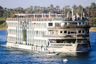 A large, multi-deck river cruise ship with ornate decorations is sailing on a wide river. The ship has several balconies, large windows, and greenery adorning the top deck. On the top deck, people are visible, enjoying the view. The riverbank is lush with tall palm trees, indicating a tropical or subtropical location. A small rowboat with two individuals can be seen in the foreground, suggesting a contrast between modern leisure and traditional transport.
