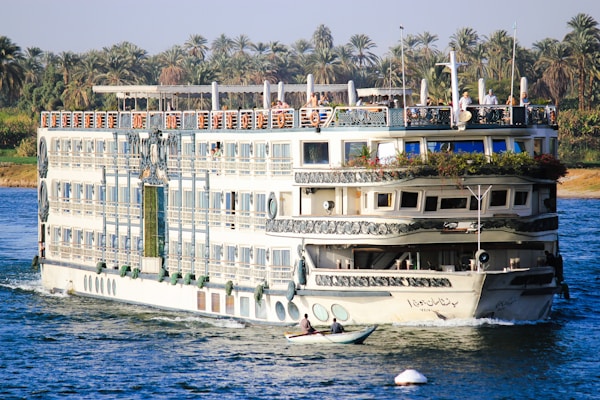 A large, multi-deck river cruise ship with ornate decorations is sailing on a wide river. The ship has several balconies, large windows, and greenery adorning the top deck. On the top deck, people are visible, enjoying the view. The riverbank is lush with tall palm trees, indicating a tropical or subtropical location. A small rowboat with two individuals can be seen in the foreground, suggesting a contrast between modern leisure and traditional transport.
