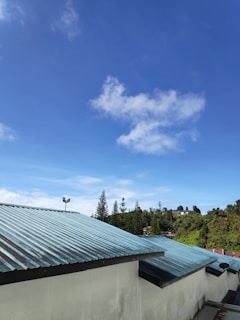 A view of a blue sky scattered with a few white clouds, above green trees and rooftops with metallic sheets. The scene is bright and clear, with tall trees in the distance and a security camera mounted on one of the rooftops.
