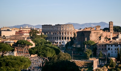 The image showcases a historic cityscape with iconic ancient architecture and surrounding greenery. In the foreground, lush trees line the streets, while the background is dominated by an ancient amphitheater with a classic Roman architectural style. There are buildings with varying historical designs spread throughout the scene, set against a backdrop of distant mountains under a clear blue sky.