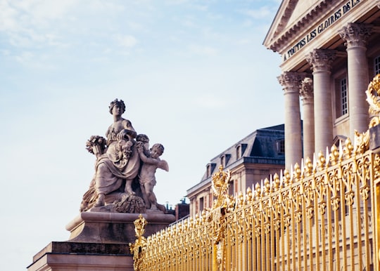 gray woman and cherubs statue in front of building with gold-colored grill fence in Versailles France