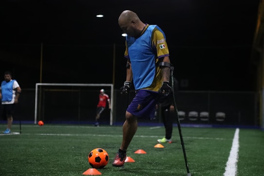 A man wearing a blue training vest is practicing on a football field. He is using crutches and moving a bright orange ball with his foot. Other people in sports attire can be seen in the background, and the field is marked with orange cones.