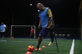 A man wearing a blue training vest is practicing on a football field. He is using crutches and moving a bright orange ball with his foot. Other people in sports attire can be seen in the background, and the field is marked with orange cones.