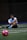 A man sits on a sports field, wearing a blue sports vest over a white shirt and black shorts. His legs are crossed, and he appears to be resting or taking a break. The background is dimly lit, with a blurred out soccer ball in the foreground and netting visible behind him.