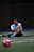 A man sits on a sports field, wearing a blue sports vest over a white shirt and black shorts. His legs are crossed, and he appears to be resting or taking a break. The background is dimly lit, with a blurred out soccer ball in the foreground and netting visible behind him.